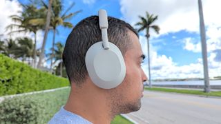 best headphone deals, Man Sony WH-1000XM5 wireless headphones outside with blue skies and palm trees in the background